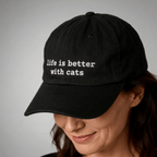 Woman wearing a black embroidered cap with the phrase life is better with cats