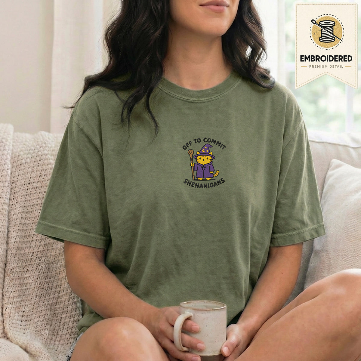 Woman wearing a moss green embroidered funny cat t shirt with Commit Shenanigans cat design while sitting indoors