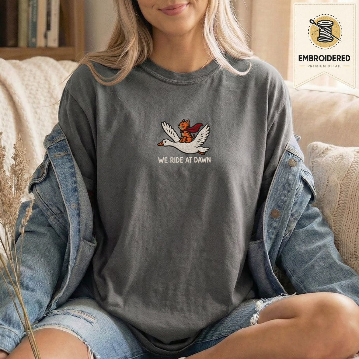 Woman wearing a pepper gray embroidered funny cat t shirt with We Ride At Dawn cat design while sitting indoors in a denim jacket