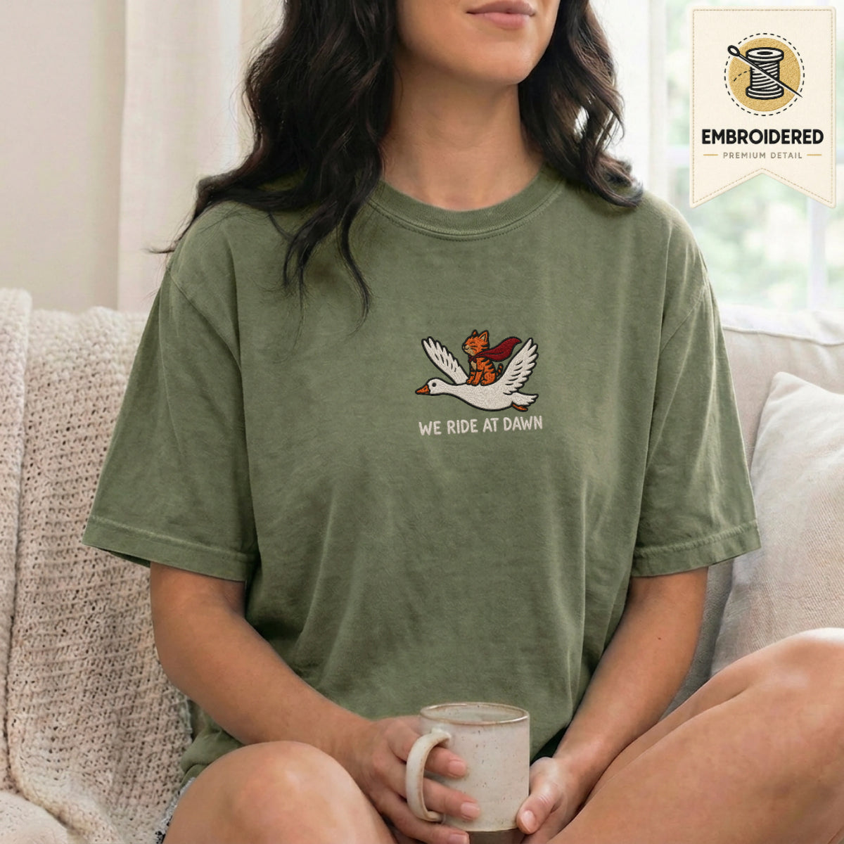 Woman wearing a moss green embroidered funny cat t shirt with We Ride At Dawn cat design while sitting indoors