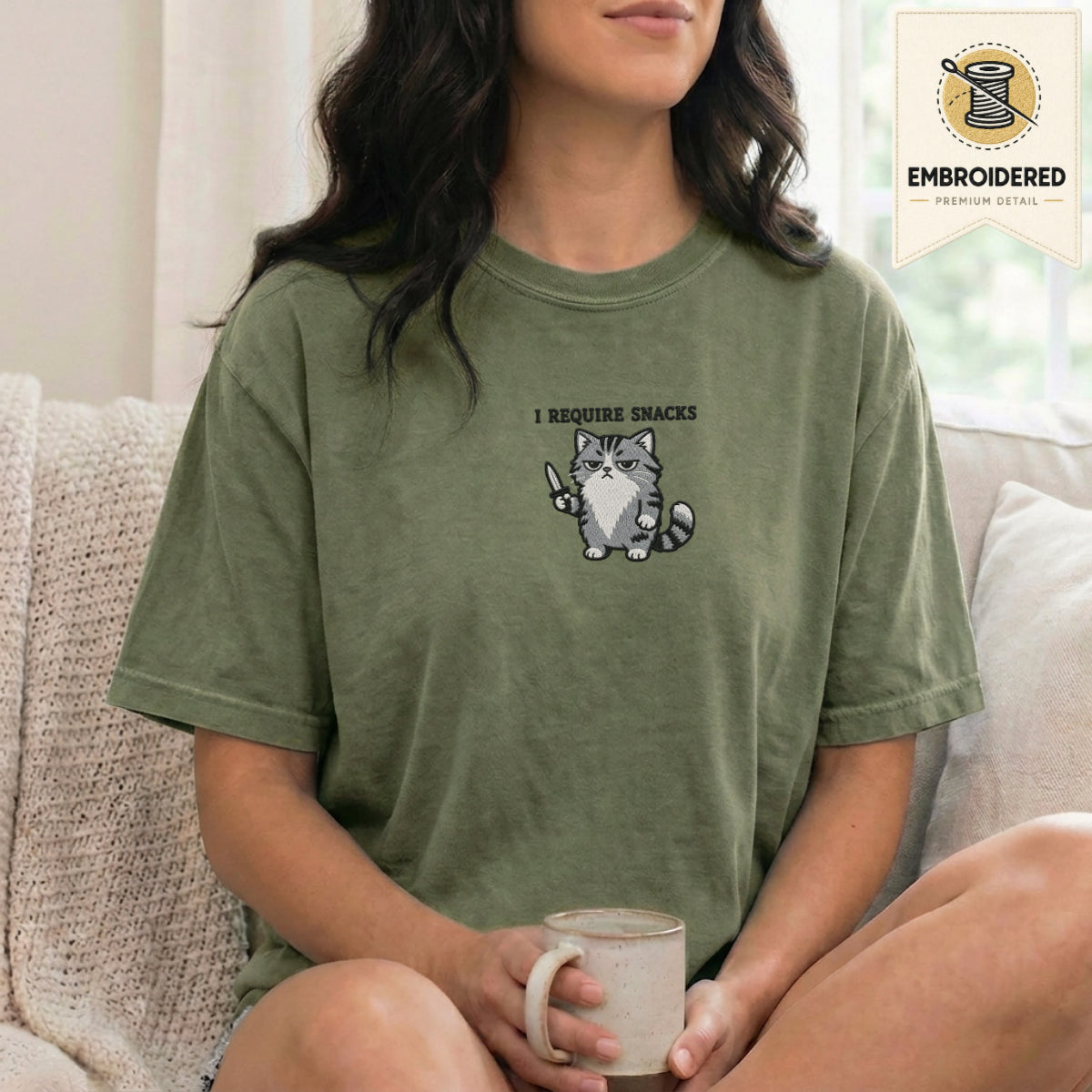 Woman wearing a moss green embroidered funny cat t shirt with Require Snacks cat design while sitting indoors