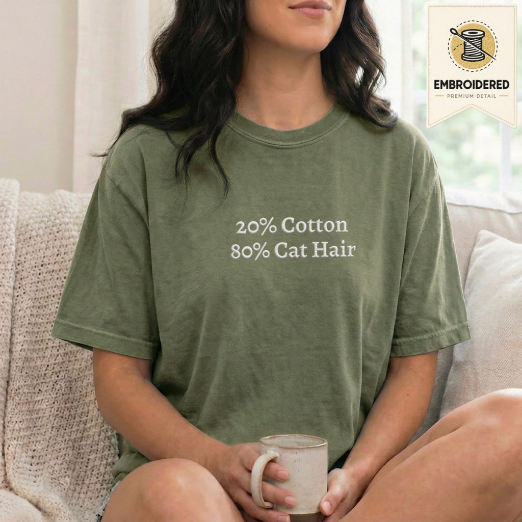Woman wearing a moss green embroidered funny cat t shirt with 20% Cotton 80% Cat Hair text while sitting indoors