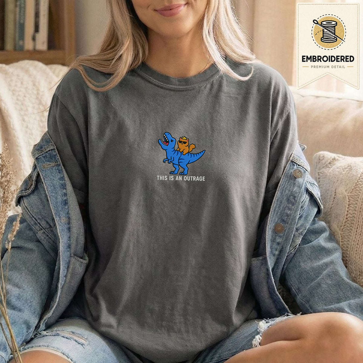 Woman wearing a pepper gray embroidered funny cat t shirt with This Is An Outrage cat design while sitting indoors in a denim jacket