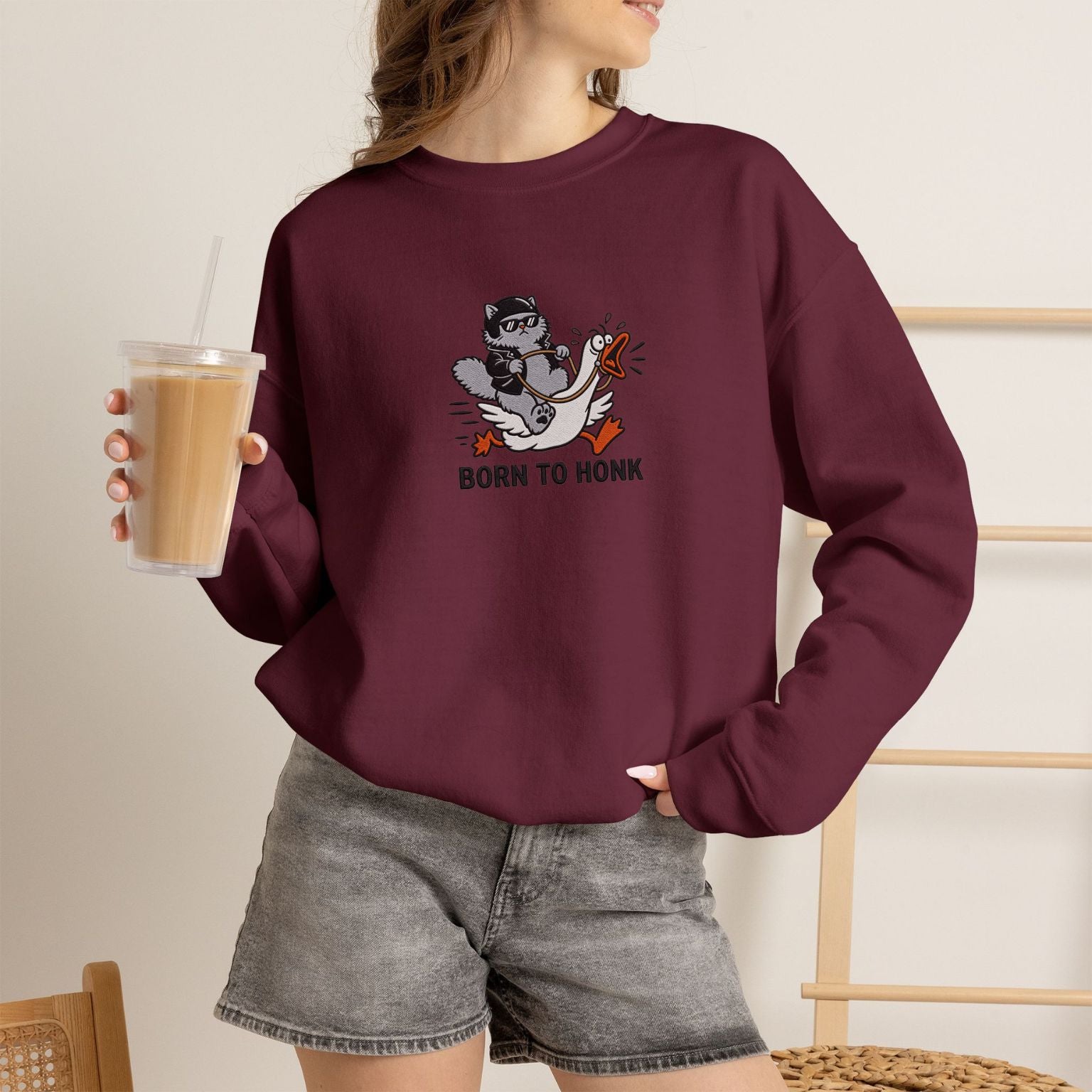 Model wearing maroon Born to Honk embroidered cat sweatshirt, holding iced coffee