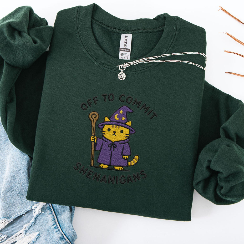Forest green embroidered cat sweatshirt with orange tabby in purple wizard robe and star hat holding a staff, Off to Commit Shenanigans text, flat lay