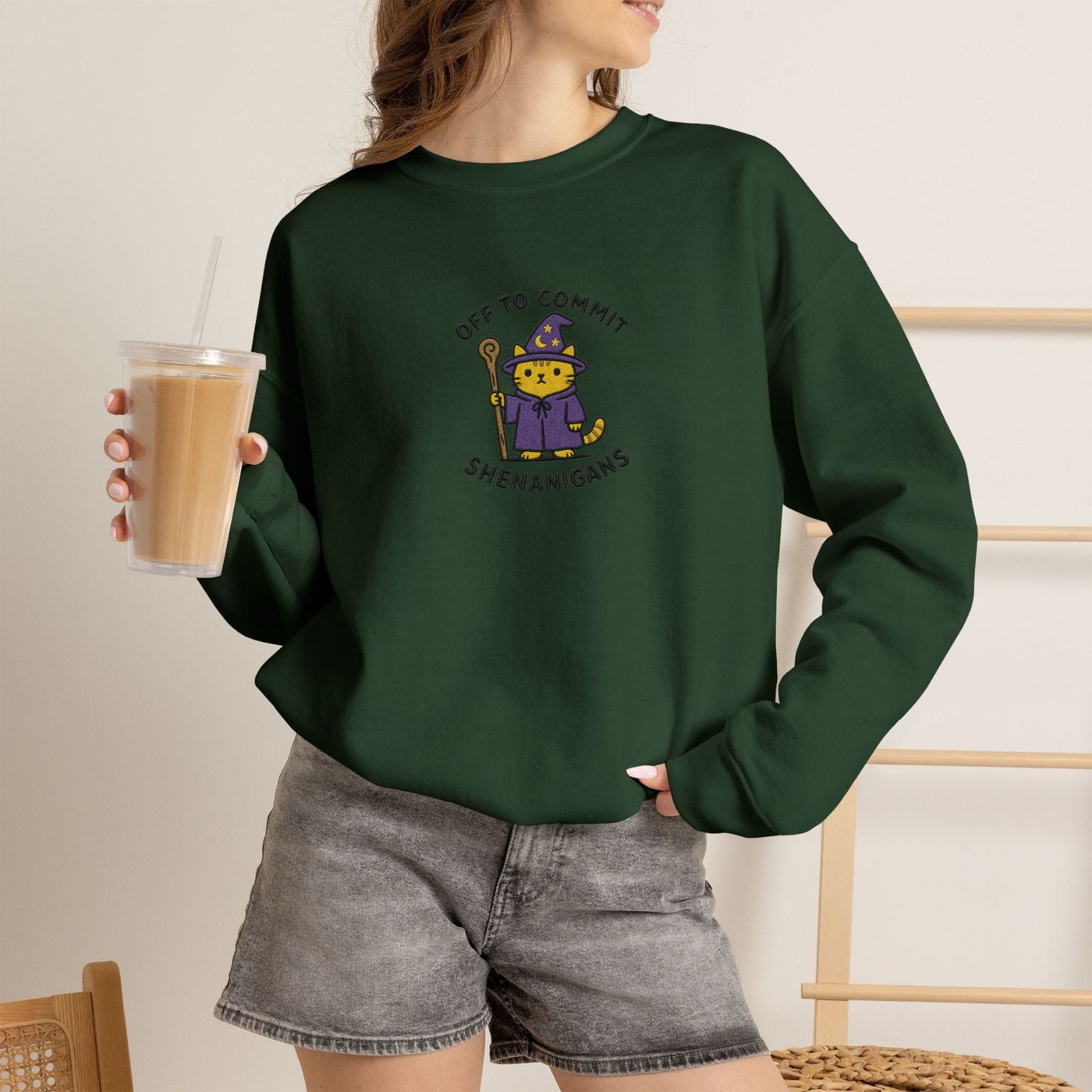 Model wearing forest green Commit Shenanigans embroidered cat sweatshirt, holding iced coffee