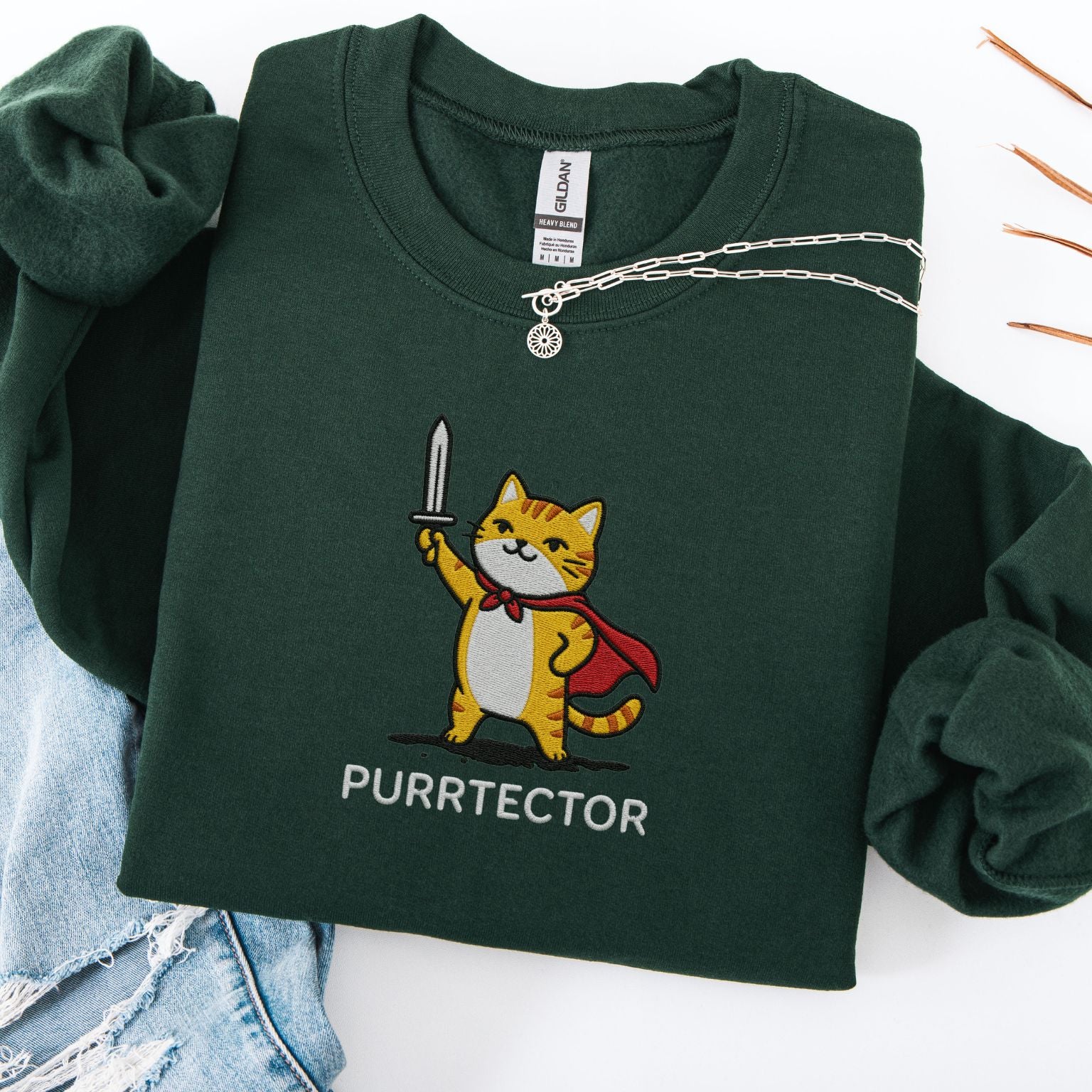 Forest green embroidered cat sweatshirt with heroic orange tabby in red cape holding sword aloft, Purrtector text, flat lay