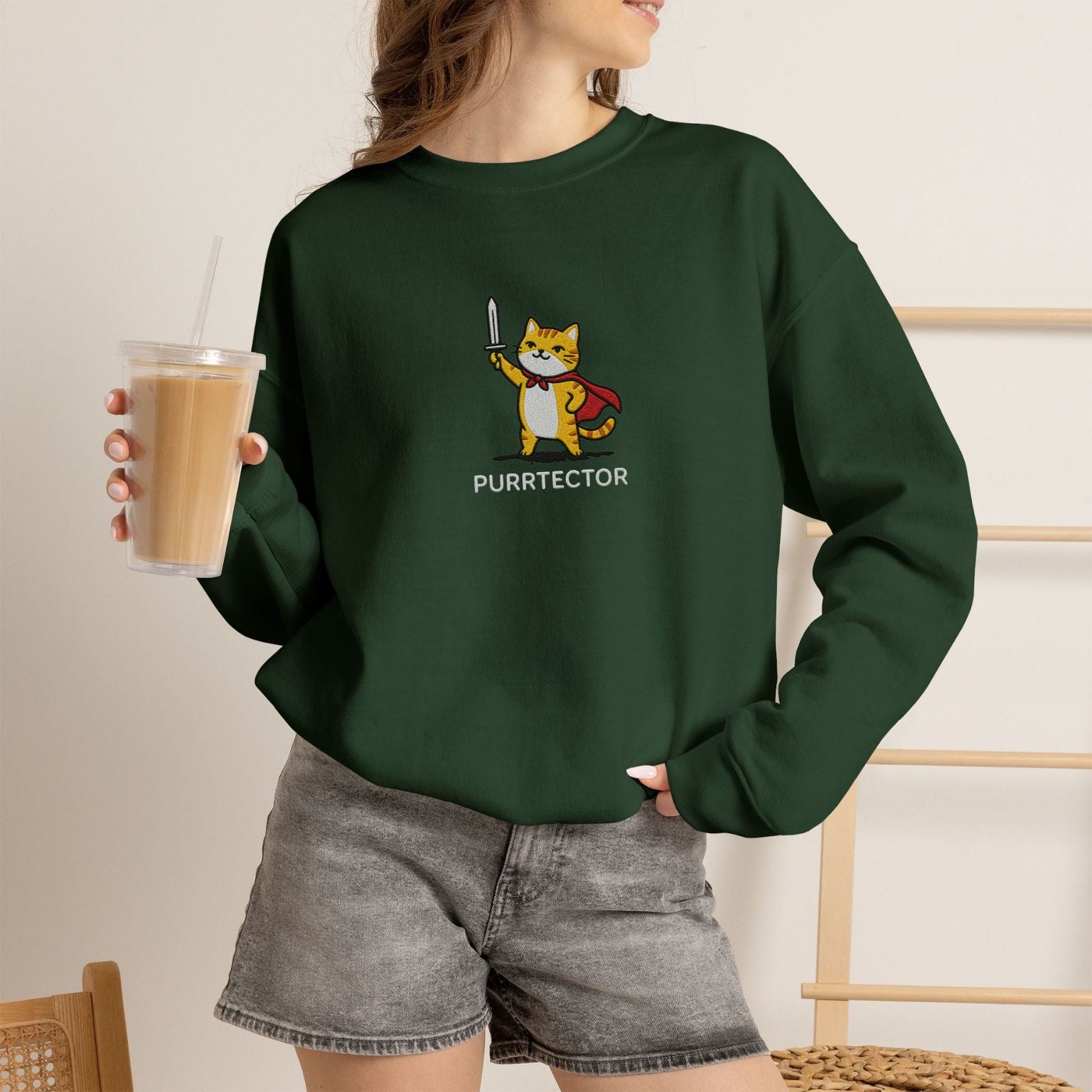 Model wearing forest green Purrtector embroidered cat sweatshirt, holding iced coffee