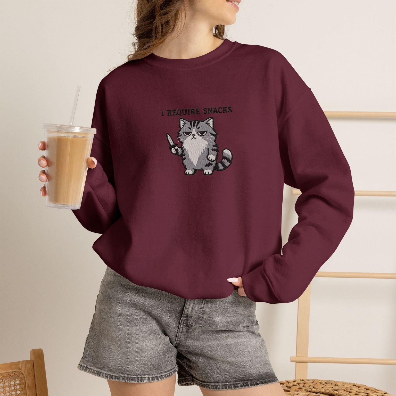 Model wearing maroon I Require Snacks embroidered cat sweatshirt, holding iced coffee