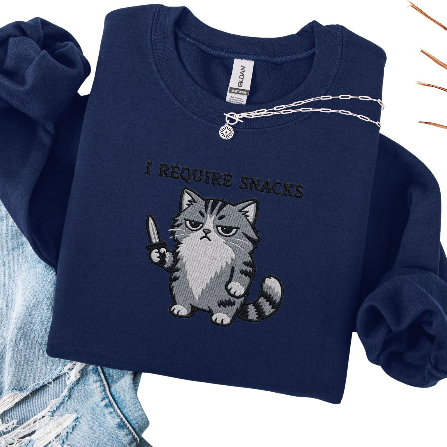 Forest green embroidered cat sweatshirt with grumpy grey cat holding a knife, I Require Snacks text, flat lay