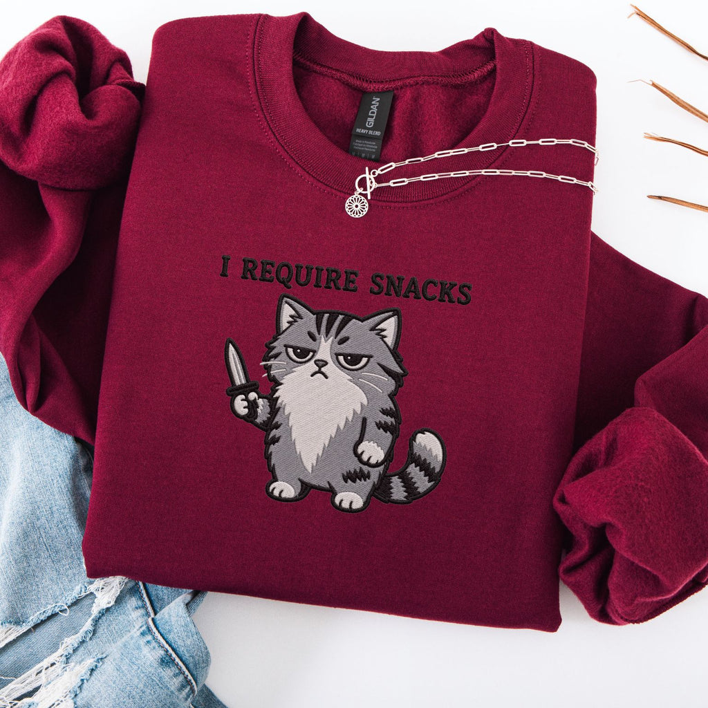Maroon embroidered cat sweatshirt with grumpy fluffy grey cat holding a knife, I Require Snacks text above, flat lay