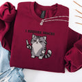 Maroon embroidered cat sweatshirt with grumpy fluffy grey cat holding a knife, I Require Snacks text above, flat lay