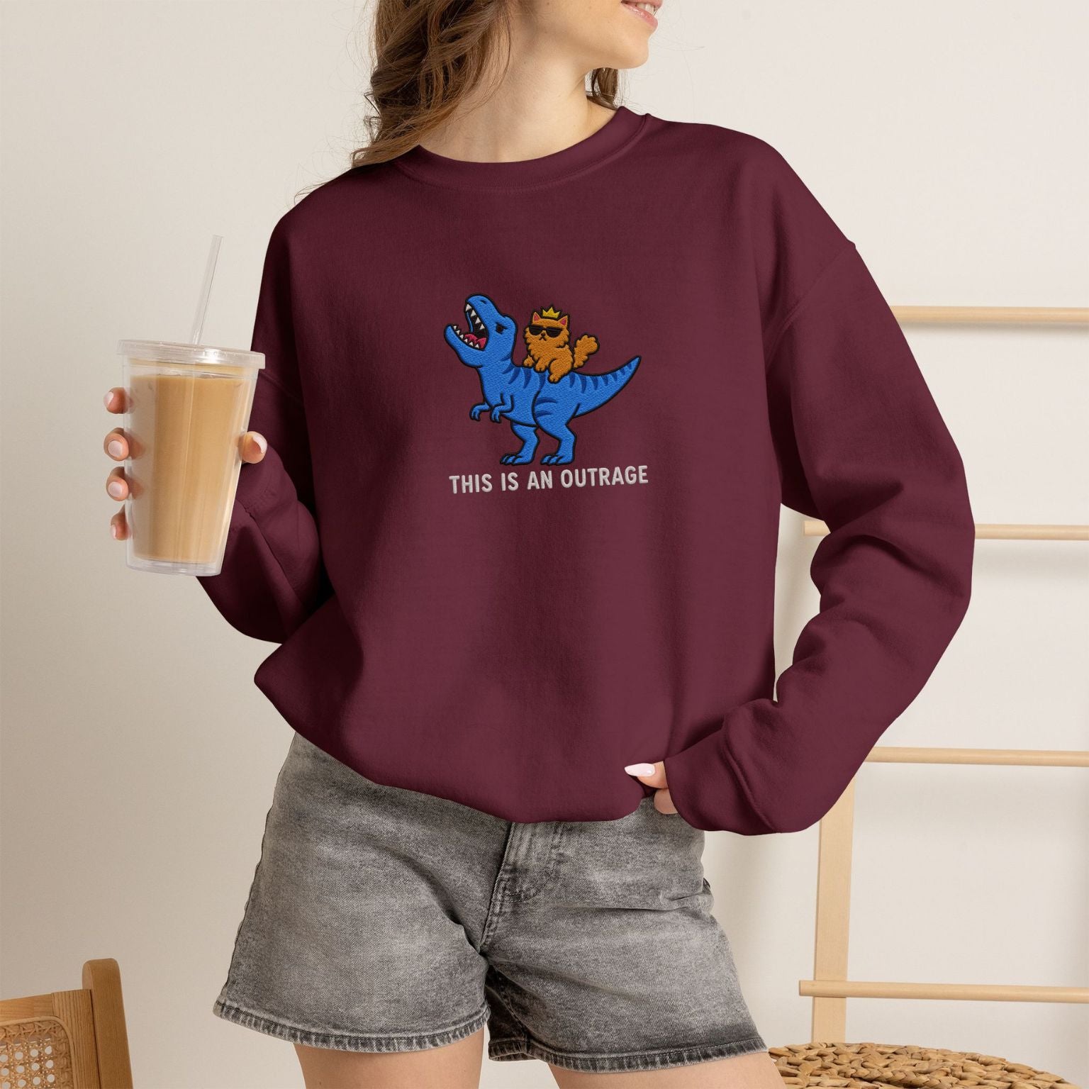 Model wearing maroon This Is An Outrage embroidered cat sweatshirt, holding iced coffee