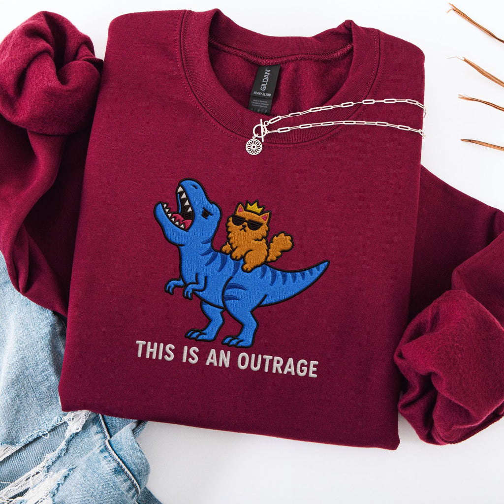 Maroon embroidered cat sweatshirt with crowned orange cat in sunglasses riding a blue T-Rex, This Is An Outrage text, flat lay