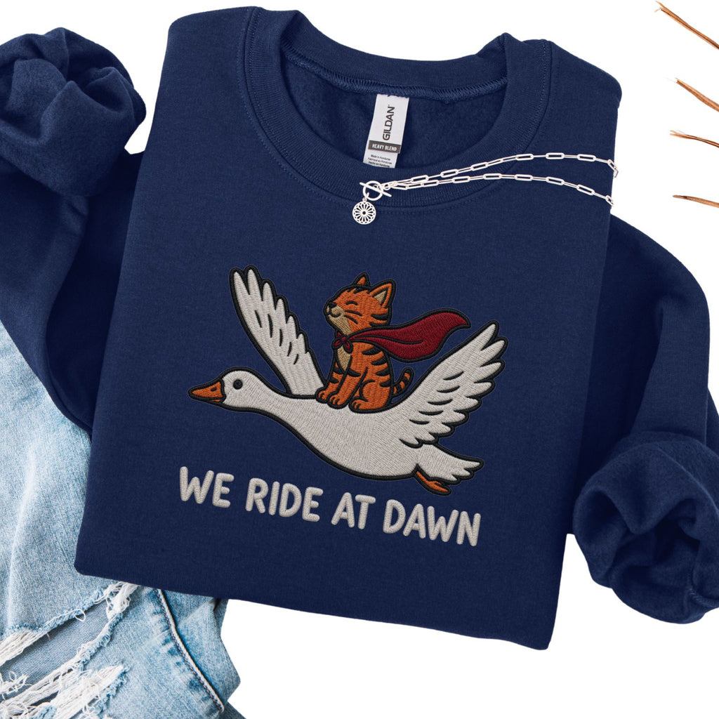 Navy embroidered cat sweatshirt — caped orange tabby riding a goose, We Ride At Dawn text, flat lay