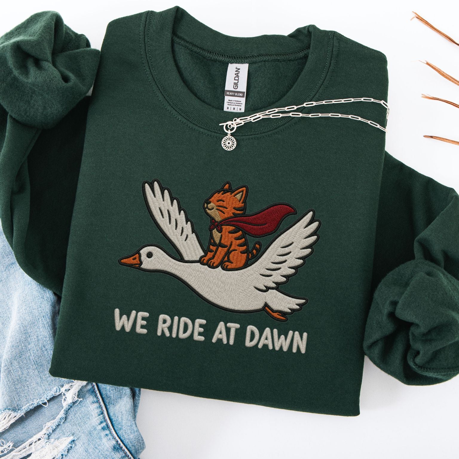 Forest green embroidered cat sweatshirt — caped orange tabby in red cape riding a white goose, We Ride At Dawn text, flat lay