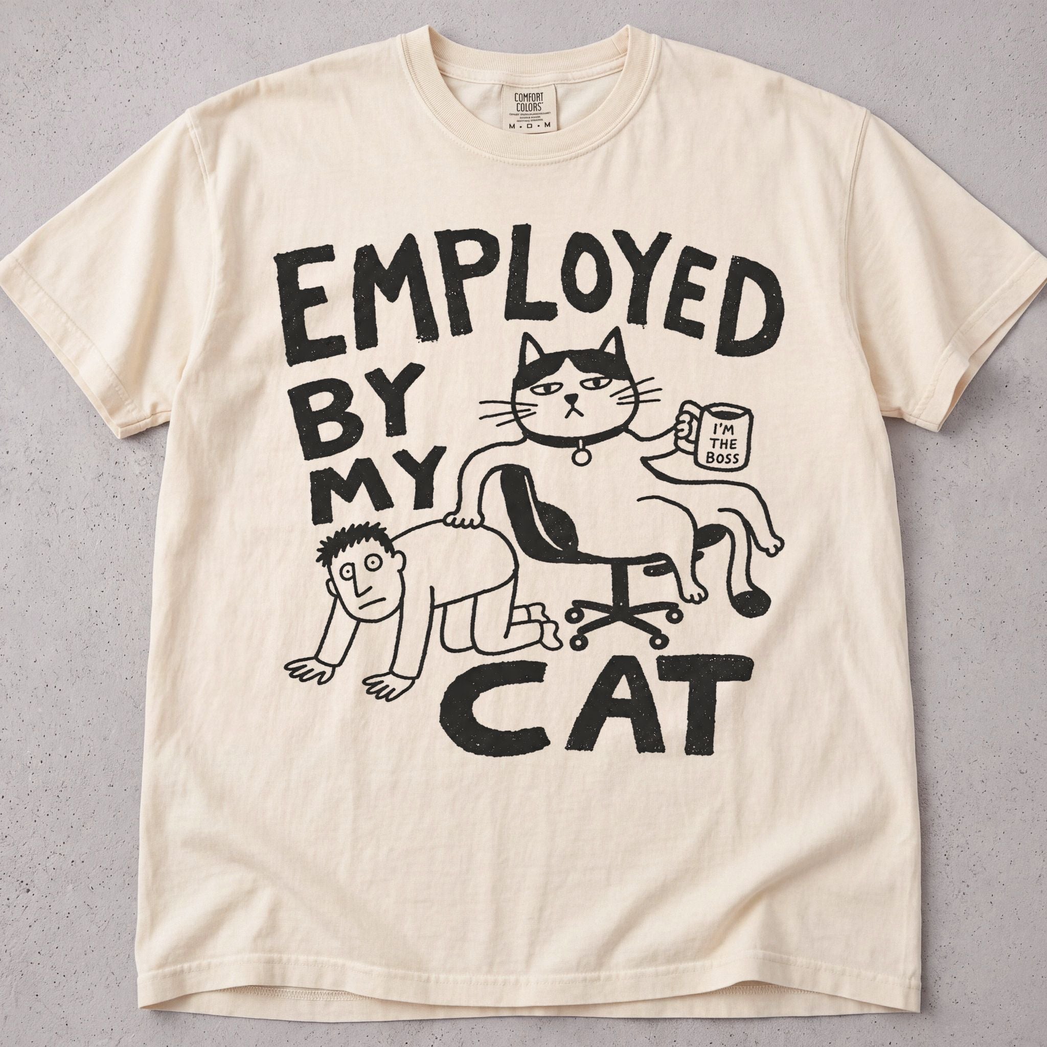 Ivory cat t-shirt with an Employed By My Cat illustration of a cat with a coffee mug, funny cat lover tee on soft Comfort Colors cotton, flat lay front view