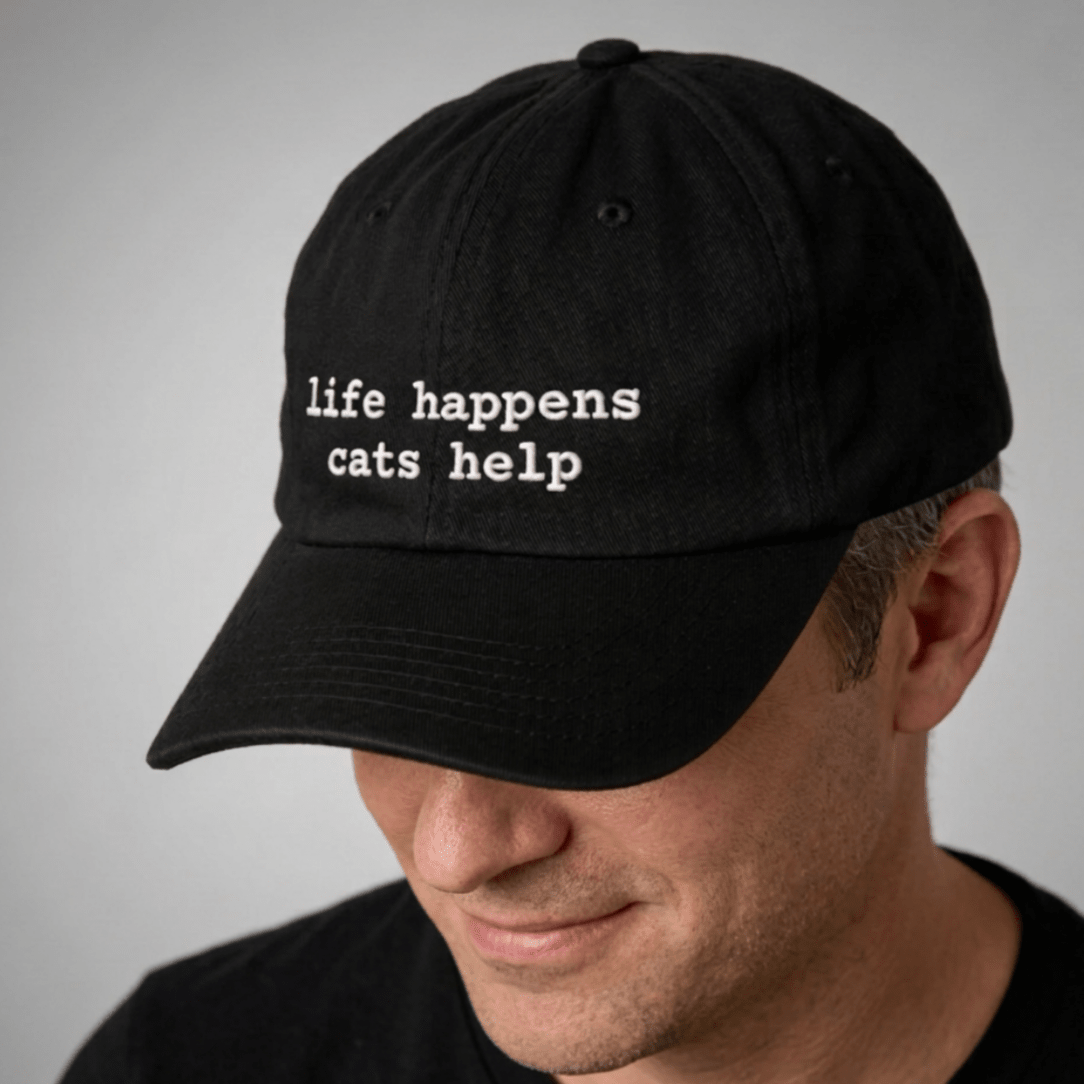 Black cap with 'life happens cats help' text worn by a person on a gray background