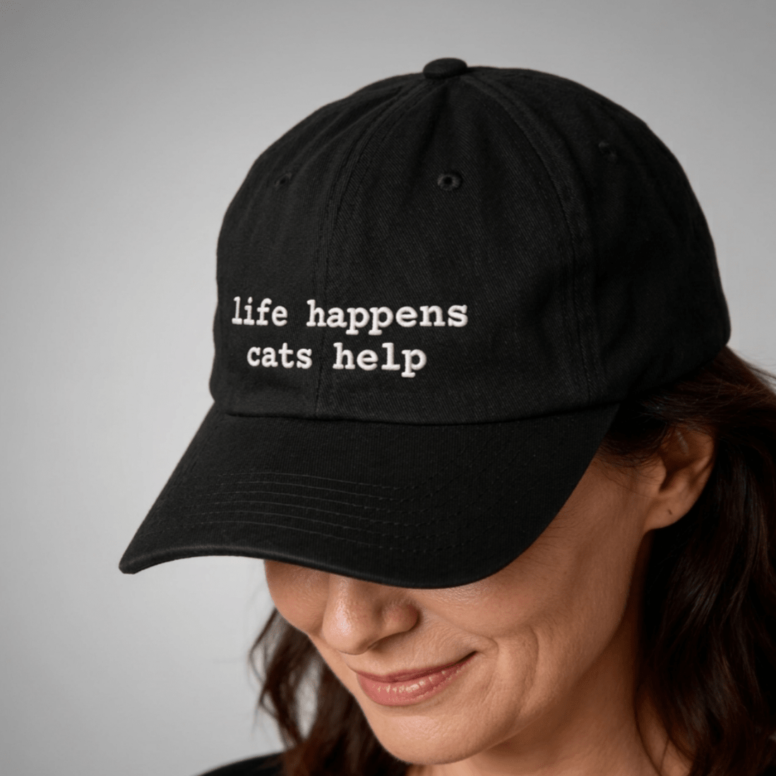 Black cap with 'life happens, cats help' text worn by a person on a gray background