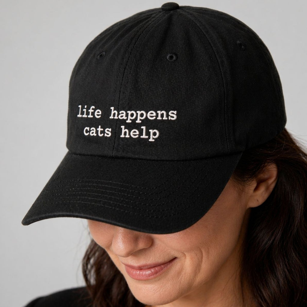 Black cap with 'life happens, cats help' text worn by a person on a gray background