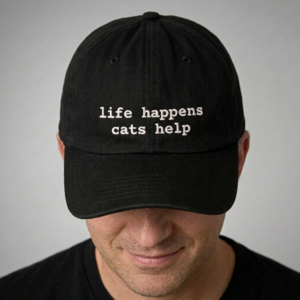 Black cap with 'life happens cats help' text worn by a person on a gray background