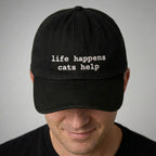 Black cap with 'life happens cats help' text worn by a person on a gray background